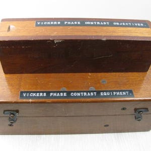 Może przedstawiać: Dwie prostokątne drewniane skrzynki do przechowywania ustawione jedna na drugiej. Górna skrzynka ma napis VICKERS PHASE CONTRAST OBJECTIVES. Dolna skrzynka posiada dwa metalowe zatrzaski oraz napis VICKERS PHASE CONTRAST EQUIPMENT. Obie skrzynki są brązowe.