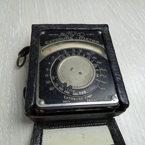 Puede incluir: Un exposímetro AVO vintage alojado en un estuche de cuero negro. El dispositivo cuenta con un dial circular plateado con marcas numéricas negras, incluyendo aperturas y tiempos de exposición, y el texto AVO EXPOSURE METER en la cara metálica.