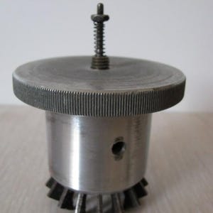 Peut inclure: Un composant de machine cylindrique en métal doté d'un dessus plat et circulaire avec un bord moleté et une tige filetée centrale avec un petit ressort. La base du composant présente une série d'ailettes métalliques rayonnantes et un petit trou circulaire est visible sur le côté.