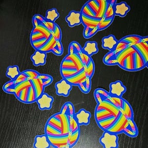 Könnte beinhalten: Eine Sammlung farbenfroher Aufkleber mit einem Planeten-Design. Jeder Aufkleber hat einen blauen Umriss, einen Regenbogen-farbenen Garnball, einen geflochtenen Garnring und drei gelbe Sternakzente. Die Aufkleber sind auf einer dunklen Oberfläche angeordnet.