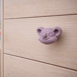 Op de afbeelding: Een lichtpaarse koala-vormige ladeknop met een gefacetteerde, geometrische oppervlaktestructuur. De knop is gemonteerd op een lichtgekleurd houten ladefront, met een gladde afwerking en horizontale houtnerfpatronen over het oppervlak.
