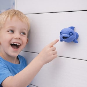 May include: A matte blue dolphin-shaped drawer knob with a smooth, rounded finish and a stylised, friendly facial expression, featuring a dorsal fin and side fins, mounted onto a white horizontal panelled surface for furniture application.