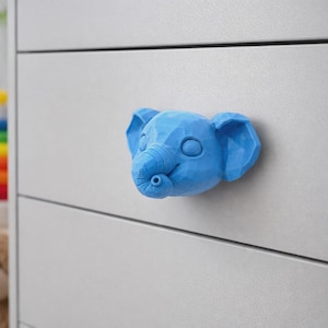 May include: A bright blue, low-poly geometric elephant head drawer knob attached to a light grey cabinet drawer. The elephant features a faceted, angular design with prominent ears, a trunk, and closed eyes, providing a textured surface.