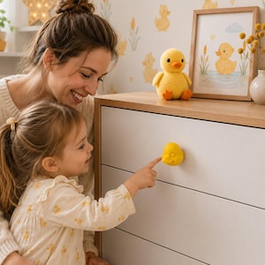 May include: A yellow duck-shaped drawer knob attached to a white dresser drawer. The knob features a three-dimensional duck head design with a small orange beak and eyes, providing a functional handle for opening the furniture drawer.