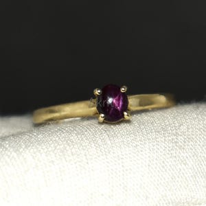 May include: A gold ring with a deep purple oval gemstone. The ring's band is polished gold, and the gemstone is held by four prongs. The ring is displayed on a white surface against a dark background.