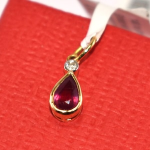 Könnte beinhalten: Ein goldener Anhänger mit einem tropfenförmigen, tiefroten Edelstein. Über dem Edelstein ist ein kleiner, runder Diamant gesetzt. Der Anhänger ist an einem weißen Band befestigt und liegt auf einer roten Oberfläche. Schmuck.
