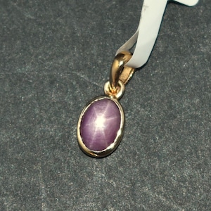 May include: A gold pendant featuring an oval, star-shaped, purple gemstone. The pendant is suspended from a white tag with text including "14K st ru" and "AKJ". The gemstone is set in a gold bezel.