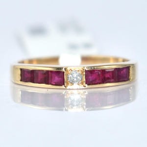 Op de afbeelding: Een gouden ring met een rij vierkant geslepen, dieprode edelstenen, met een enkele, kleine, vierkante diamant in het midden. De ring wordt getoond op een reflecterend oppervlak.