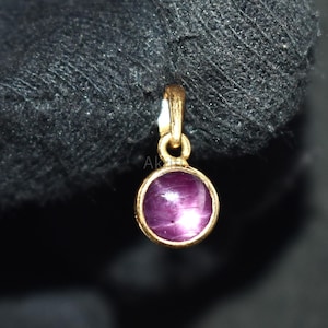 May include: A small, round pendant with a gold-coloured setting. The pendant features a deep pink gemstone with a star-like effect. The gemstone is set in a simple, elegant gold frame. The pendant is suspended from a small loop.