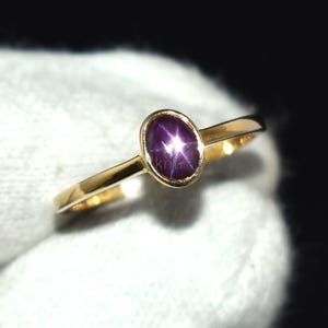 Può includere: Un anello d'oro con una gemma ovale viola scuro con un motivo a forma di stella. La fascia dell'anello è sottile e lucida, e la gemma è incastonata in una semplice lunetta. L'anello è mostrato su sfondo bianco.
