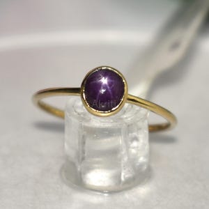 Op de afbeelding: Een gouden ring met een ronde, dieppaarse edelsteen met een stervormig patroon. De edelsteen is gezet in een gouden bezel en de ring heeft een fijne gouden band. De ring wordt gepresenteerd op een heldere, vierkante standaard.