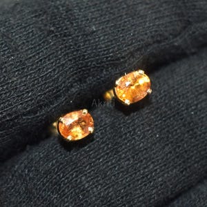 Könnte beinhalten: Ein Paar goldene Ohrstecker, jeweils mit einem oval geschliffenen, orangefarbenen Edelstein. Die Ohrringe sind in einer einfachen Goldfassung gefasst, die die leuchtende Farbe der Steine hervorhebt. Der Hintergrund ist ein dunkler, strukturierter Stoff.