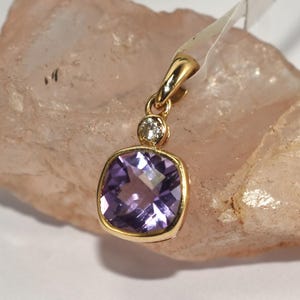 May include: A gold pendant featuring a square, purple gemstone and a small, round diamond. The pendant is suspended from a gold bail. The pendant is displayed on a light pink, textured surface.