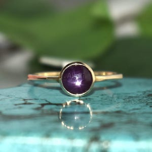 May include: A gold ring featuring a circular, deep purple gemstone. The ring rests on a turquoise surface, with blurred green leaves in the background. The gemstone is the central element, highlighting its colour and form.