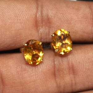 May include: A pair of oval-shaped, golden-yellow gemstone stud earrings. The gemstones are set in a gold-coloured metal. The earrings are displayed on a hand against a dark background.