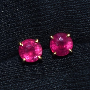 Pode incluir: Um par de brincos de rubi. Cada brinco apresenta uma pedra preciosa redonda rosa vibrante em uma configuração de quatro garras douradas. Os brincos são exibidos em um fundo escuro e texturizado.