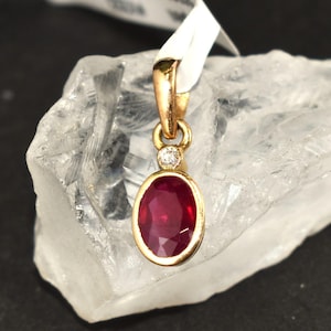 May include: A gold pendant featuring an oval, deep red gemstone and a small diamond. The pendant hangs from a gold bail. The pendant is displayed on a clear, crystalline surface.