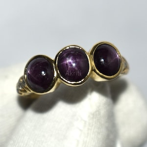 May include: A gold ring featuring three oval, dark purple gemstones. The gemstones have a star-like effect. The ring is held by a gloved hand against a white background. The word "Akriti" is visible on the ring.