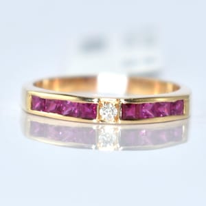 Puede incluir: Un anillo de oro con una fila de piedras preciosas rojas oscuras de corte cuadrado y un pequeño diamante. La banda de oro pulido del anillo sujeta las piedras preciosas al ras. El anillo se muestra sobre una superficie reflectante.