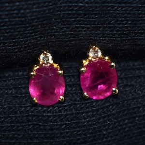 Könnte beinhalten: Ein Paar goldene Ohrstecker, jeweils mit einem leuchtend rosa, oval geschliffenen Edelstein. Ein kleiner, klarer Akzentstein sitzt auf jedem rosa Edelstein. Die Ohrringe sind vor einem dunkelblauen Hintergrund platziert.