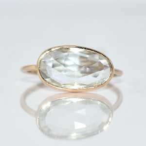 以下が含まれることがあります： 大きな淡い緑色の楕円形の宝石をあしらったゴールドリング。 宝石はシンプルなゴールドバンドにセットされており、ミニマルでエレガントなデザインになっています。 リングは反射する表面に表示されています。