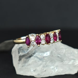 以下が含まれることがあります： ゴールドリング。マーキス型のルビーとラウンドダイヤモンドが交互に並んでいます。リングは、暗い背景に対して透明な結晶質の表面にセットされています。宝石は鮮やかな赤と白です。