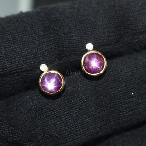 Op de afbeelding: Een paar oorstekers met een gouden zetting. Elke oorbel heeft een ronde, dieppaarse edelsteen met een sterren-effect en een klein, helder edelsteenaccent. De oorbellen worden gepresenteerd op een zwart oppervlak.