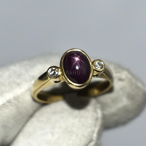 Può includere: Un anello d'oro con una grande gemma ovale rosso scuro con effetto a stella, affiancata da due gemme più piccole e trasparenti. L'anello ha una fascia d'oro, che presenta un design classico ed elegante. Questo è un gioiello.