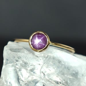 May include: A gold ring featuring a round, purplish-red star ruby gemstone. The ring is set in a gold bezel and rests on a light blue, crystalline surface against a dark background. The ring's band is thin and delicate.