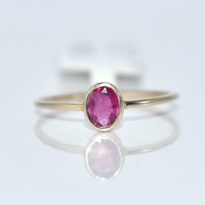 May include: A gold ring with a vibrant oval-cut ruby gemstone. The ring's band is slender and polished, and the ruby is set in a bezel setting. The ring is displayed on a reflective surface, highlighting its design.