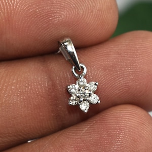 May include: A small, silver-colored pendant featuring a flower design. The flower is composed of multiple small, round, clear gemstones, likely diamonds, set in a starburst pattern. The pendant is suspended from a small loop.