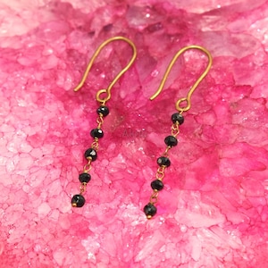 May include: A pair of gold-colored dangle earrings with black beads. The earrings are hanging against a pink background. The earrings have a simple design, with a hook at the top and a chain of beads.