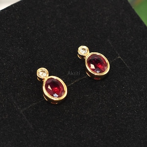 Peut inclure: Une paire de boucles d'oreilles à clous en or avec des pierres précieuses ovales rouge foncé. Chaque boucle d'oreille a une petite pierre précieuse ronde et transparente au-dessus de la pierre ovale. Les boucles d'oreilles sont présentées sur une surface noire.