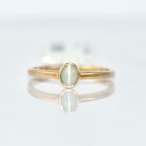 May include: A gold ring with a light green oval gemstone. The ring has a thin band and a simple, elegant design. The gemstone is set in a classic bezel setting, and the ring is displayed on a reflective surface.