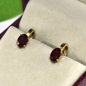 May include: Two gold pendant earrings with oval, deep red gemstones, presented in a burgundy jewelry box with a cream-colored interior. The earrings are displayed in a close-up shot, highlighting their design and color.