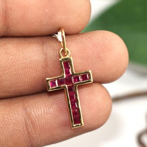 Puede incluir: Colgante dorado en forma de cruz, con incrustaciones de piedras preciosas rojas rectangulares. La cruz está suspendida de un pequeño lazo dorado en la parte superior.