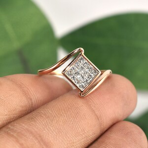 Puede incluir: Un anillo de oro rosa con un cuadrado de peque&ntilde;as piedras brillantes. La banda del anillo tiene un dise&ntilde;o curvo, creando un aspecto moderno y elegante. El anillo se muestra sobre un fondo verde borroso.