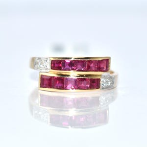 以下が含まれることがあります： ラップアラウンドデザインの2つのゴールドリング。各リングには、正方形にカットされた鮮やかな赤い宝石と、小さく透明なアクセントストーンが並んでいます。リングは白い背景に配置され、鏡面反射があります。