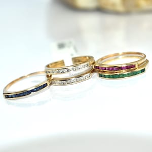 Könnte beinhalten: Vier goldene Ringe mit verschiedenen Edelsteinanordnungen. Ein Ring hat eine Reihe blauer Saphire, ein anderer klare Steine, und der dritte hat Rubine und Smaragde. Die Ringe werden auf einer weißen Oberfläche präsentiert.