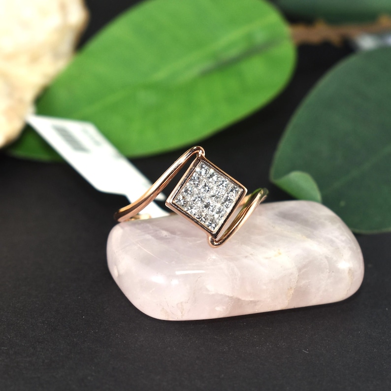 Puede incluir: Un anillo de oro rosa con un centro cuadrado con diamantes. El anillo est&aacute; colocado sobre una piedra rosa p&aacute;lido, con hojas verdes en el fondo. La banda del anillo se curva alrededor del dedo.