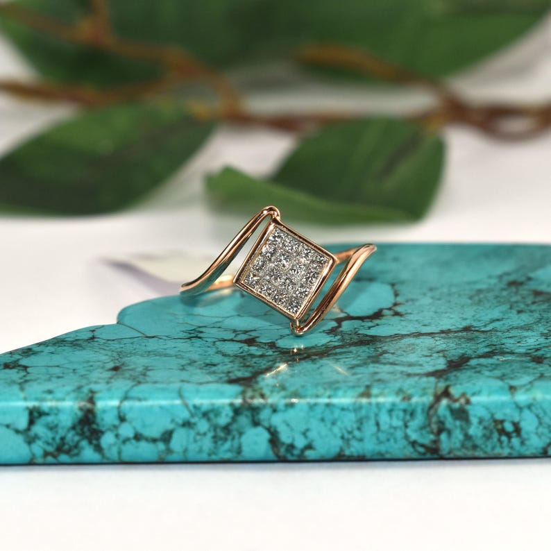 Puede incluir: Un anillo de oro rosa con un centro cuadrado con diamantes. La banda del anillo tiene un dise&ntilde;o curvo y elegante. El anillo se muestra sobre una superficie turquesa, con hojas verdes en el fondo.