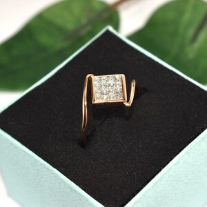 Puede incluir: Un anillo de oro rosa con un cuadrado de peque&ntilde;as piedras brillantes. La banda del anillo tiene un dise&ntilde;o curvo, y el cuadrado est&aacute; centrado. El anillo se muestra en una caja negra con interior azul claro.