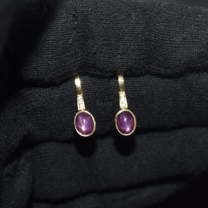 May include: A pair of gold earrings featuring oval, star-shaped, purple gemstones. Each earring has a small diamond accent and a hook closure. The earrings are presented on a black surface. The word "Akriti" is printed on the surface.