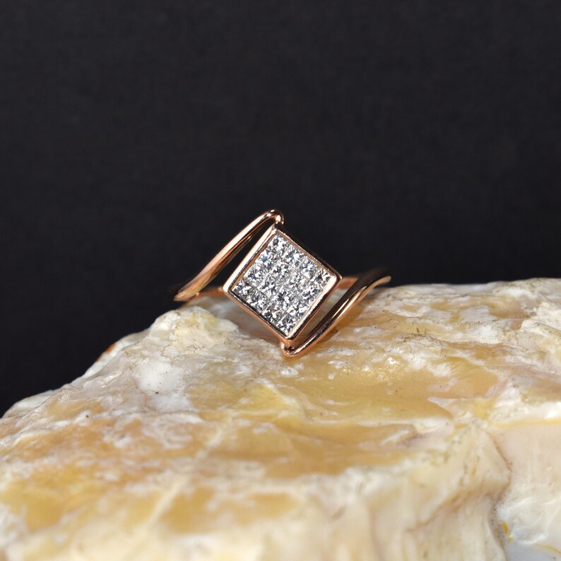 Puede incluir: Anillo de oro rosa con un cuadrado de peque&ntilde;as piedras claras. La banda del anillo tiene un dise&ntilde;o curvo, con el engaste cuadrado en el centro. El anillo se muestra sobre una superficie clara y texturizada sobre un fondo oscuro.