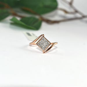 Puede incluir: Anillo de oro rosa con un grupo cuadrado de peque&ntilde;as piedras preciosas transparentes. El anillo tiene un dise&ntilde;o &uacute;nico con una banda curva que fluye hacia el engaste cuadrado. El anillo est&aacute; sobre un fondo blanco con una hoja y una rama verdes borrosas en el fondo.