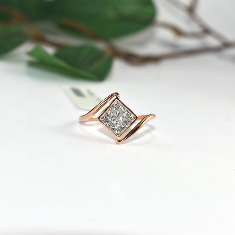 Puede incluir: Anillo de oro rosa con un grupo cuadrado de peque&ntilde;as piedras preciosas transparentes. El anillo tiene un dise&ntilde;o &uacute;nico con una banda curva que fluye hacia el engaste cuadrado. El anillo est&aacute; sobre un fondo blanco con una hoja y una rama verdes borrosas en el fondo.