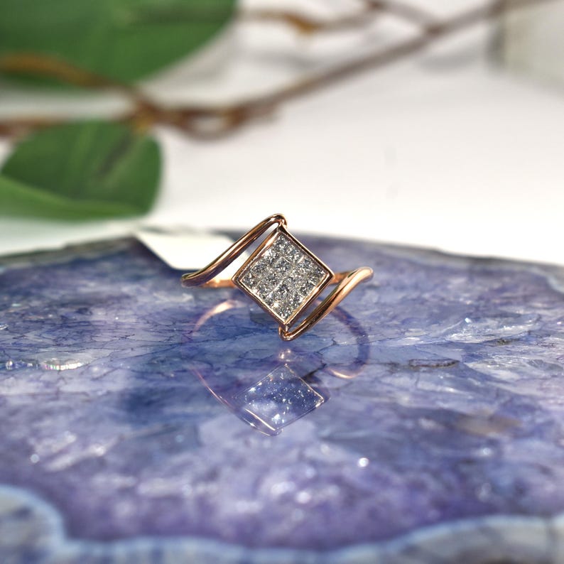 Puede incluir: Un anillo de oro rosa con un grupo cuadrado de peque&ntilde;as piedras transparentes en el centro. El anillo tiene un dise&ntilde;o &uacute;nico y fluido con bandas curvas que se extienden desde el engaste cuadrado. Se muestra sobre una superficie azul y morada.