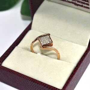 Puede incluir: Un anillo de oro rosa con un grupo cuadrado de peque&ntilde;as piedras preciosas transparentes. El anillo se muestra en un joyero burdeos con un interior color crema. El dise&ntilde;o del anillo presenta una banda curva &uacute;nica.