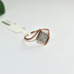 Puede incluir: Anillo de oro rosa con un centro cuadrado con diamantes. El anillo presenta un dise&ntilde;o &uacute;nico y asim&eacute;trico con una banda curva. Los diamantes est&aacute;n engastados en un patr&oacute;n cuadrado, creando un efecto brillante. El anillo se muestra sobre una superficie blanca.