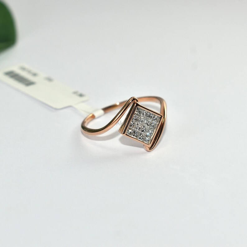 Puede incluir: Anillo de oro rosa con un centro cuadrado con diamantes. El anillo presenta un dise&ntilde;o &uacute;nico y asim&eacute;trico con una banda curva. Los diamantes est&aacute;n engastados en un patr&oacute;n cuadrado, creando un efecto brillante. El anillo se muestra sobre una superficie blanca.